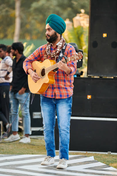 молодой индийский сикх в бирюзовом головном уборе, красной рубашке и синих джинсах играет на гитаре на открытом воздухе - new delhi audio стоковые фото и изображения