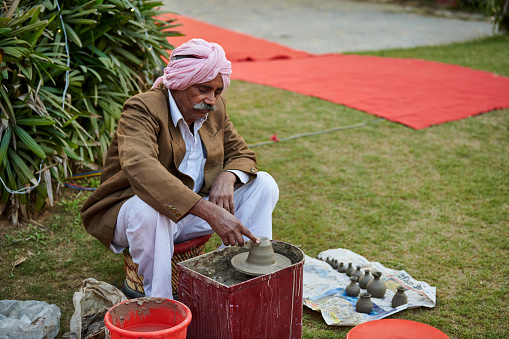 New Delhi, India - 10.12.2022 - Outdoor public park. Old indian sikh man in pink pagri headwear, white pants and brown jacket makes pottery items from clay, old potter creates clay vases on potters wheel
