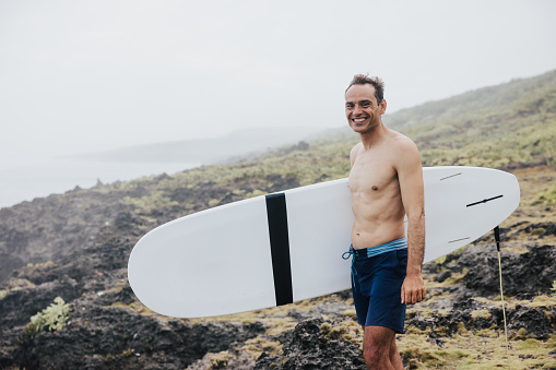 A surfer, unfazed by the cloudy weather, navigates the rocky shoreline, his surfboard under his arm, ready for a adventure.