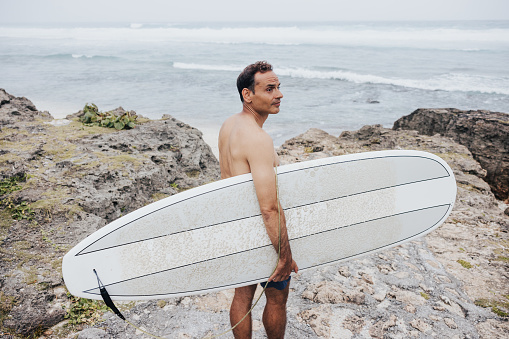 On a misty day at the rocky beach, a surfer with a surfboard makes his way towards the ocean waves at Okinawa