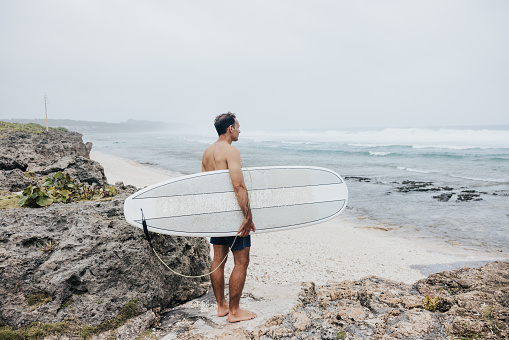 On a misty day at the rocky beach, a surfer with a surfboard makes his way towards the ocean waves at Okinawa