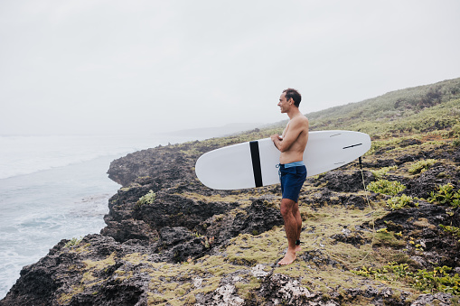 A surfer, unfazed by the cloudy weather, navigates the rocky shoreline, his surfboard under his arm, ready for a adventure.