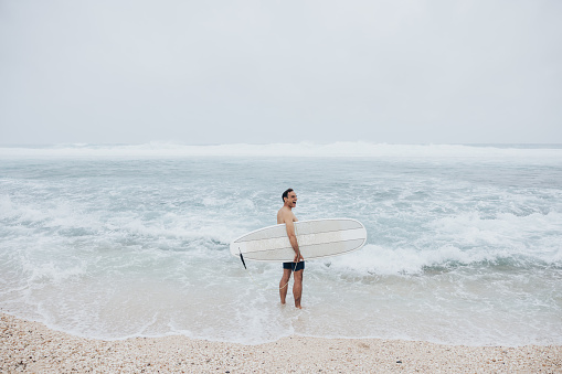 With a radiant smile, the surfer stands in the refreshing water, his surfboard ready for the next adventure.