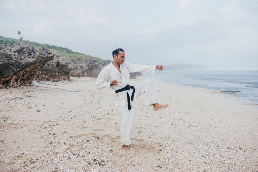 A martial artist, dressed in traditional white gi, executes a precise karate kata on the sandy beach with the ocean as a stunning backdrop