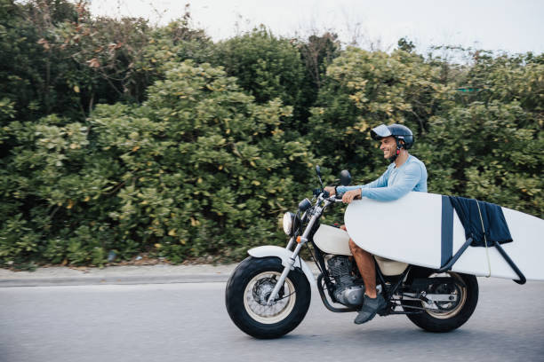 Cruising along the coastal road, a surfer balances his surfboard securely on his motorcycle, headed for the waves.
