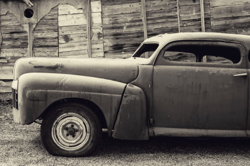 A customized vintage coupe sits outside an old garage.  Toned black and white image.