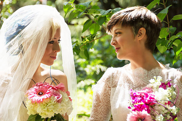Horizontal outdoor garden shot of two young women in wedding dresses smiling at each other.