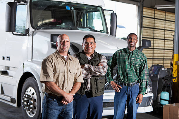 Multi-ethnic men standing next to semi-truck Three truck drivers standing in front of the cab of a white semi-truck parked in the garage of a warehouse.  The group of multracial men are standing in a row, smiling at the camera, looking relaxed and confident.  They are wearing blue jeans and button-down shirts.  The man in the middle is Hispanic. trucking photos stock pictures, royalty-free photos & images
