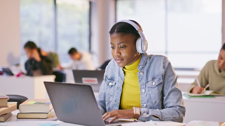 Headphones, computer and black woman in a class listening to music, playlist or album at university. Technology, laptop and African student working on assignment streaming song or radio in college.