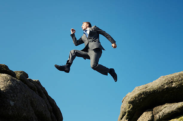 Man in suit jumping between towering rocks Young businessman in midair between two boulders with a blue sky above him.  He wears a grey suit and blue tie.  His arms are extended. leap of faith stock pictures, royalty-free photos & images