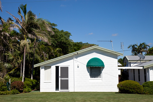 Quanit tidy little white cottage house with only one front window – landscaped with green grass and beautiful blue sky – Queensland Australia. Click to see more...