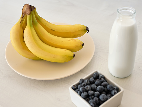 Bunch of fresh bananas on a plate with blueberries and a bottle of milk on white wooden kitchen table.