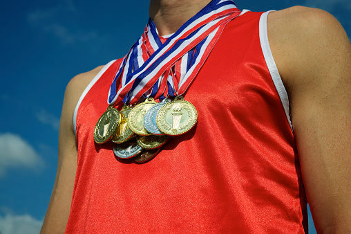 Athlete in red athletic shirt stands with gold, silver, and bronze medals in front of bright blue sky