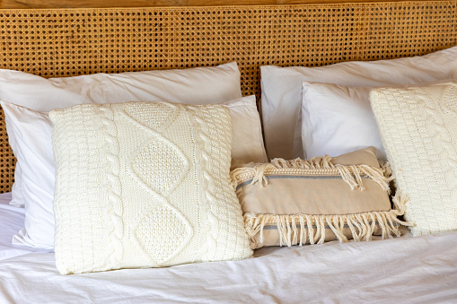 An inviting bedroom setup featuring white bedding accented with cream colored knitted and tasseled pillows. A unique rattan patterned headboard completes the serene ambiance