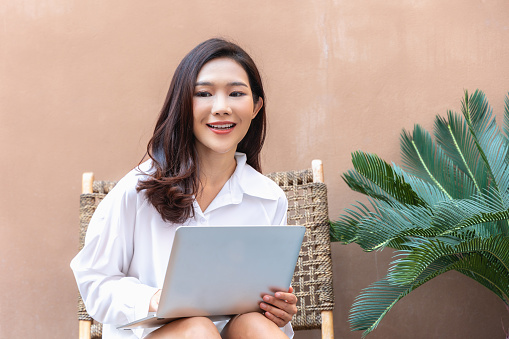 In a cozy urban corner, a radiant Asian woman confidently engages with her laptop. The synergy of modern tech and natural greenery encapsulates the essence of today's flexible work culture