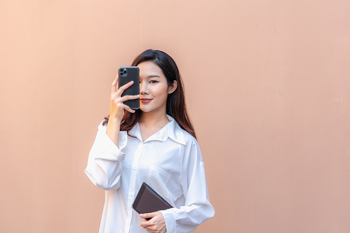 In an urban setting, a stylish asian woman effortlessly captures a moment with her smartphone. Her poised demeanor combines with chic attire, embodying the essence of modern elegance