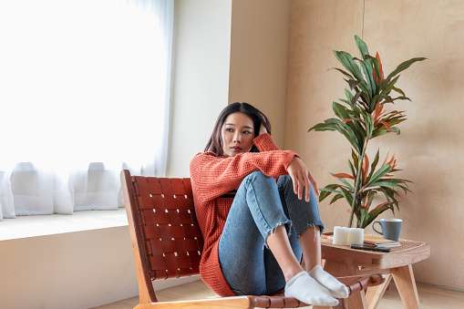 A contemplative woman sits by the window, draped in a vibrant knit sweater. Sunlit room accents, a wooden table with candles, and a tall indoor plant complete the peaceful ambiance