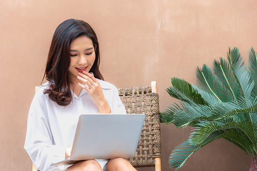 In a cozy urban corner, a radiant Asian woman confidently engages with her laptop. The synergy of modern tech and natural greenery encapsulates the essence of today's flexible work culture