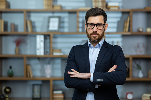 Portrait of mature experienced male boss, businessman with crossed arms seriously thinking looking at camera, senior investor in business suit inside office workplace wearing glasses.