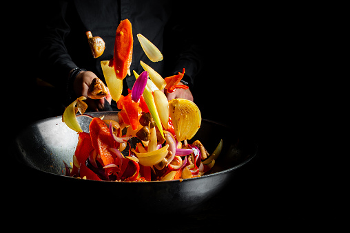 Closeup of chef throwing vegetable mix from wok pan on black background.