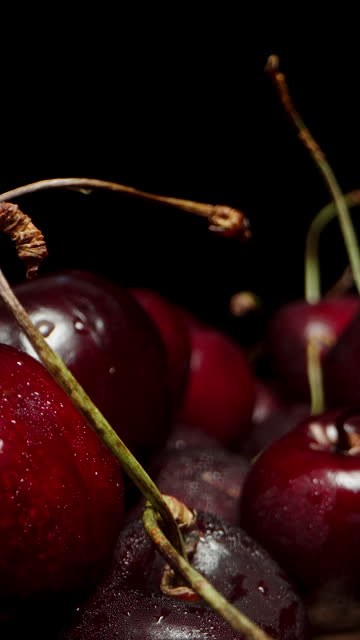Vertical video. A large quantity of cherries emerges from darkness into the light. Close-up shot. Sloder
