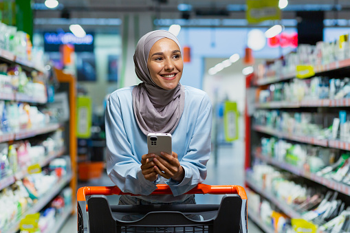 Shopping. A young beautiful Muslim woman in a hijab walks and chooses between the shelves with goods in a supermarket with a trolley. He is holding a phone with a list, application, online payment.