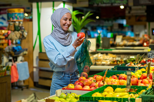 Portrait of a female buyer in a supermarket, a Muslim woman in a hijab chooses an apple fruit, happy with a large assortment of products and smiles, woman shopping in a large grocery store.