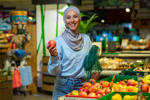 Portrait of a female buyer in a supermarket, a Muslim woman in a hijab is smiling happy and looking at the camera, choosing apples and fruits in a large grocery store.