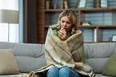 Single mature woman sitting at home in the living room on the sofa, wrapped in a blanket trying to keep warm, missing and broken heating in the house
