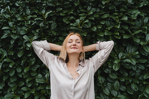 Portrait of relaxed woman with eyes closed on background of green leaves wall. Concept of outside of office, Work-Life-Balance, Taking Break. Zen and balance people. Stability through mental health.