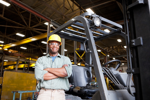 African American worker (30s) standing by forklift in factory.