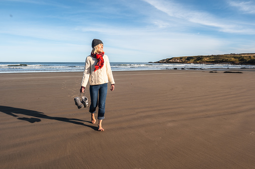 Senior woman walking along the waterline on the beach at Sandend, Moray, Scotland.