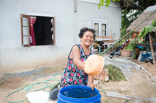 Mature thai farm woman is watering and cleaning in front of her house and home in small village Parangmee in district Noen Maprang in province of Phitsanulok. Woman is purring water with a bowl out of a barrel in her garden area. Captured from rural street through village
