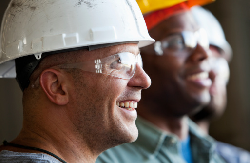 Close up of group of multi-ethnic construction workers wearing hard hats and safety glasses.  Focus on man in foreground (30s).