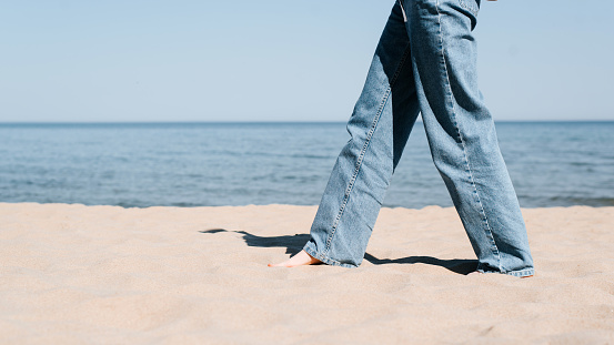 Barefoot woman walking on hot sand on the beach, close-up of female legs in blue jeans side view.