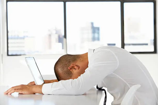 Frustrated Businessman Sitting At Desk In Office Using Laptop With Head On Table.