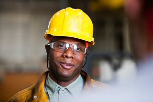 African American worker (30s) wearing hardhat and safety glasses, talking to co-worker out of focus in foreground.