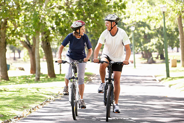 mature couple cyclisme à travers un parc - cycling senior adult sports helmet men photos et images de collection