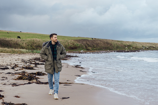 A wide shot of a young adult male walking along a beach alone at Newton-by-the-Sea in Northumberland, North East England. He is dressed in warm clothing, with his hands in his pockets, as he looks out to sea with a look of contemplation. Behind him is a view of the beach, sea and grassy headland beyond.