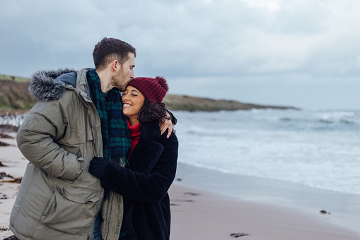 A shot of a couple embracing whilst on a beach walk at Newton-by-the-Sea in Northumberland, North East England. The male has his arm around his girlfriend and is kissing her on the head affectionately, as she embraces him with a smile and closed eyes. Behind them is a view of the beach and sea.
