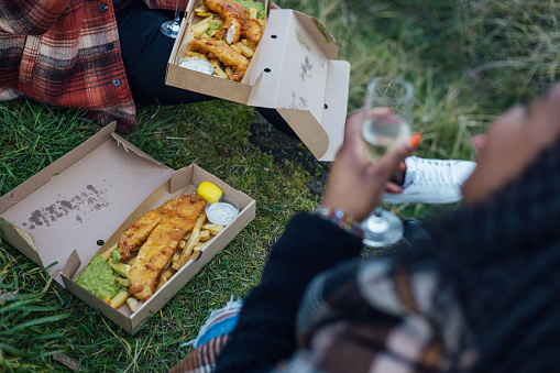 An over-the-shoulder close up shot of two unrecognisable people enjoying takeaway fish and chips outdoors in the countryside at Newton-by-the-Sea in Northumberland, North East England. The female who is blurred at the edge of the frame is holding a Champagne flute filled with a non-alcoholic beverage.