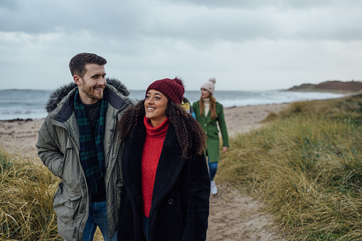 A shot of a young couple walking together ahead of their friends on a sandy path leading inland from a beach at Newton-by-the-Sea in Northumberland, North East England. They are both smiling as they walk. wearing warm clothing. Behind them is the beach and sea beyond, where waves lapping the shore are visible.