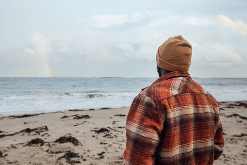 A waist-up shot of a young adult male standing on a beach at Newton-by-the-Sea in Northumberland, North East England. He is wearing warm clothing and has his back to the camera and is looking out to sea, enjoying the view. There is seaweed on the beach and waves lapping the shoreline, with a rainbow in partial view above the sea.