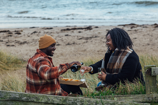 A side-view shot of a young couple sitting on the edge of a beach amongst long grass at Newton-by-the-Sea in Northumberland, North East England. They are holding take-out fish and chips, whilst the male pours non-alcoholic Prosecco into their Champagne flutes. They both have big smiles on their faces, looking at the glasses.