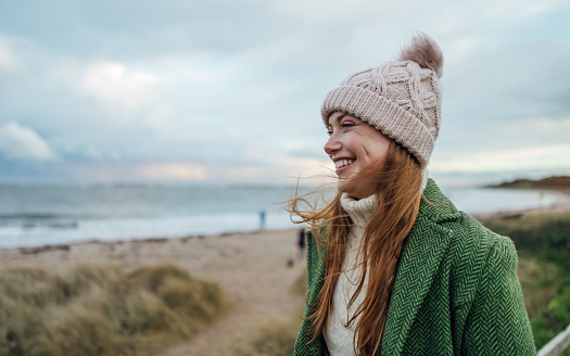 A side-view shot of a young female adult smiling whilst on a beach walk at Newton-by-the-Sea in Northumberland, North East England. She is looking straight ahead, to the left of the frame, with a wide contented smile. She is wearing a winter coat and woollen bobble hat, and behind her is a view of the beach and sea beyond.