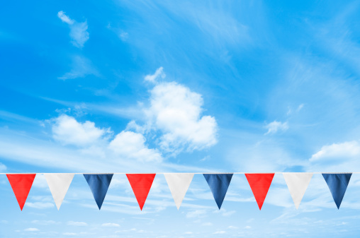 A row of red white and blue bunting hanging across a blue sky. Alternating triangles of colour are hanging from a white ribbon to form the bunting. The colours represent the British flag. They are in a line on a warm summers day with a bright blue sky behind them.