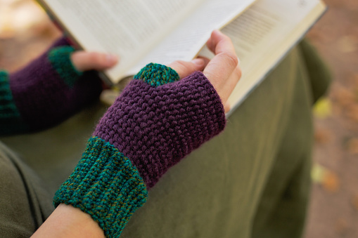The girl turns the page of the book. A woman in fingerless mittens reads a book in an autumn park. Crochet mitts made of wool and acrylic yarn. The concept of reading, relaxing with a book, walking in the park, selective focus