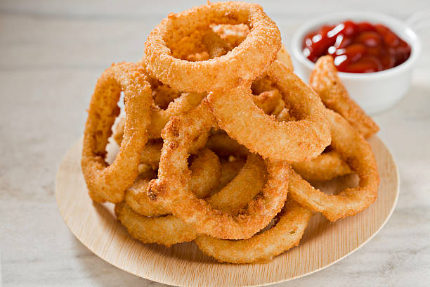 Onion Rings And Ketchup An extreme close up of a small bamboo plate with a stack of golden brown onion rings and a small container of ketchup in  the foreground. Shot on a white marble surface. fried onion rings stock pictures, royalty-free photos & images