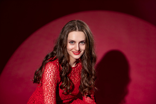 Portrait of a young woman with long brown hair in a red dress on a red background. She is smiling and looking at camera.