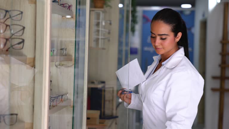 Female ophthalmologist looking at a prescription and grabbing a pair of glasses from display at an optician’s shop